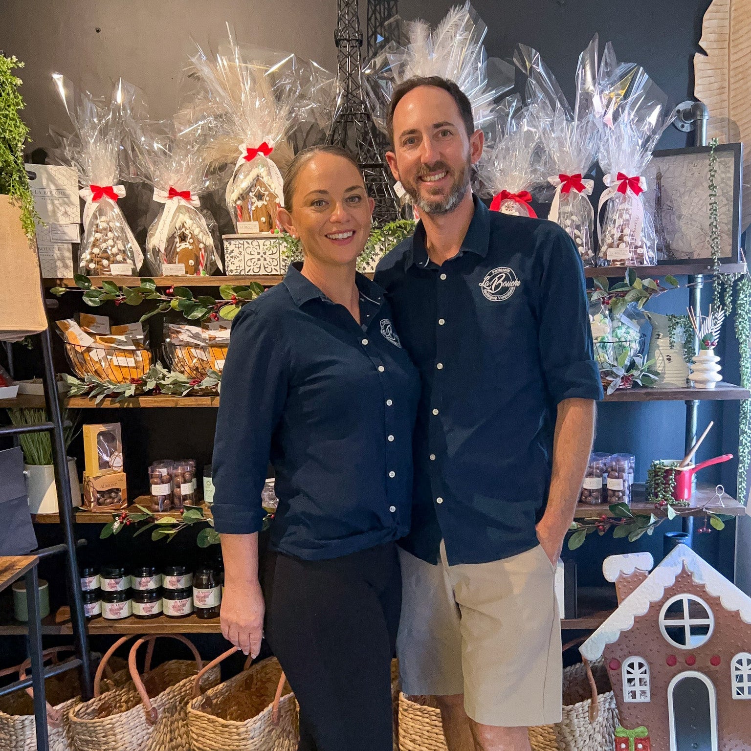 A man and a woman standing together in a gift shop or bakery setting, wearing matching navy blue button-up shirts with a circular white logo on the chest. The background is filled with shelves of wrapped gift baskets, jars, and holiday decorations.