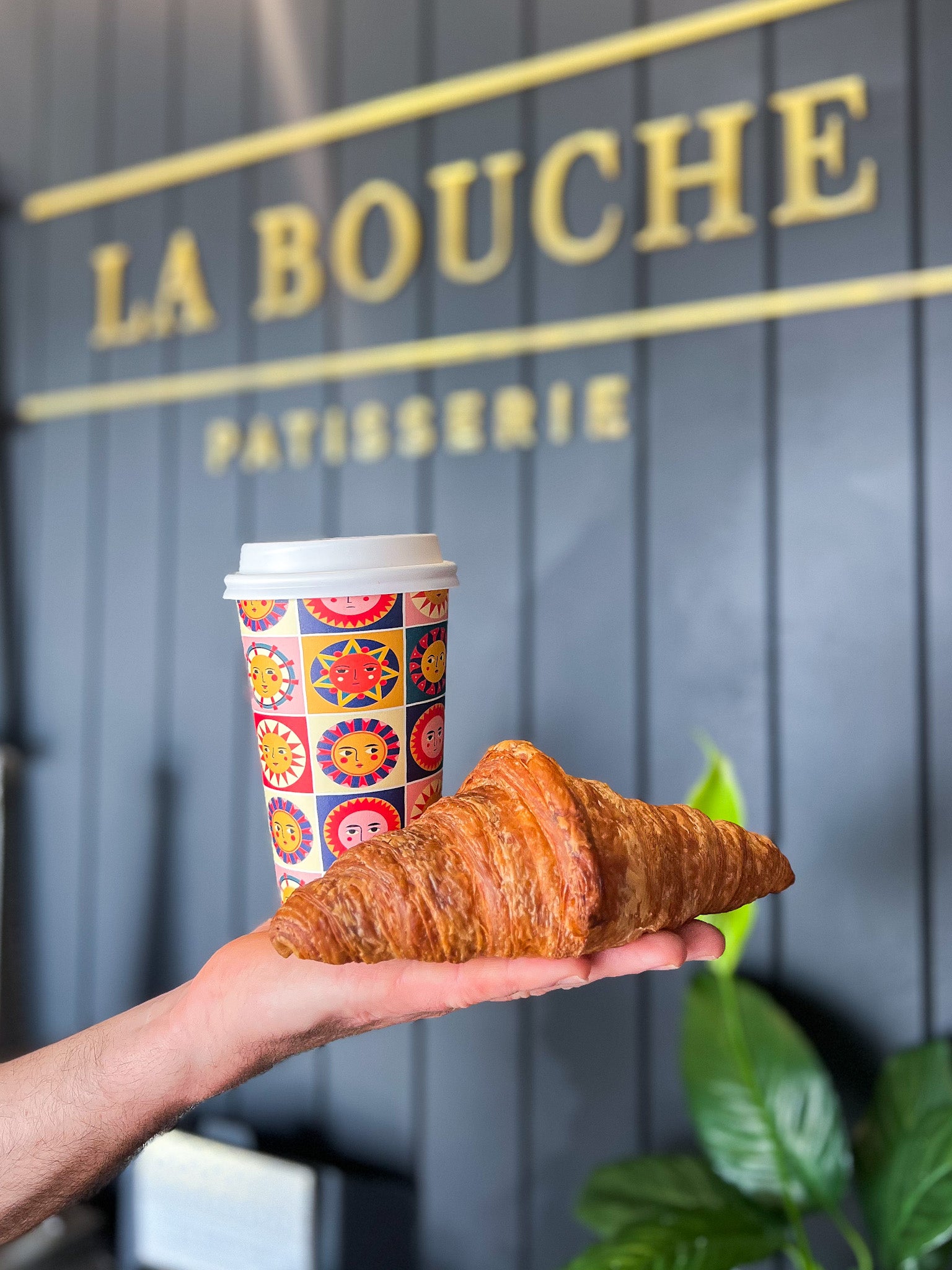 A hand holds a golden-brown croissant and a colorful paper coffee cup in front of a dark gray wall with gold 'LA BOUCHE Patisserie' signage.